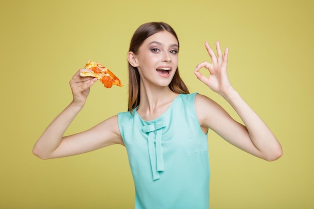노란색 배경에 아쿠아 블루 드레스에 행복 한 여자. 모델이 먹는 맛있는 피자 조각