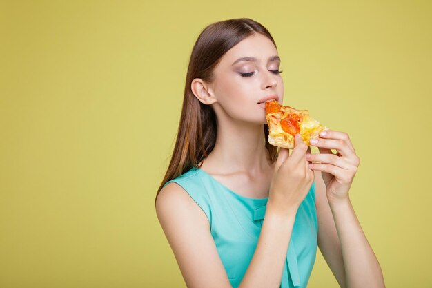 노란색 배경에 아쿠아 블루 드레스에 행복 한 여자. 모델이 먹는 맛있는 피자 조각