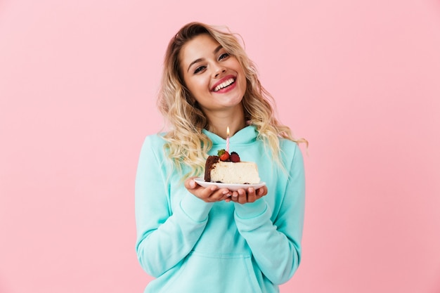 Happy woman in basic clothing holding piece of birthday cake with candle, isolated over pink wall