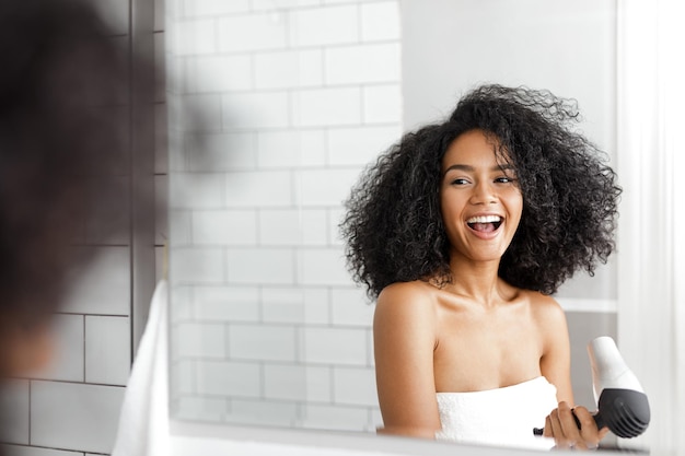 Счастливая женщина в ванной сушит свои кудрявые волосы сушилкой.