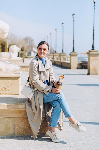 Happy woman in beige trench coat and blue jeans having breakfast on the beach. Seafront walking