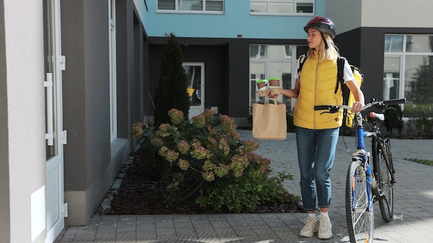 Happy woman on a bike delivering coffee and food in paper bag to customer man home Food delivery concept