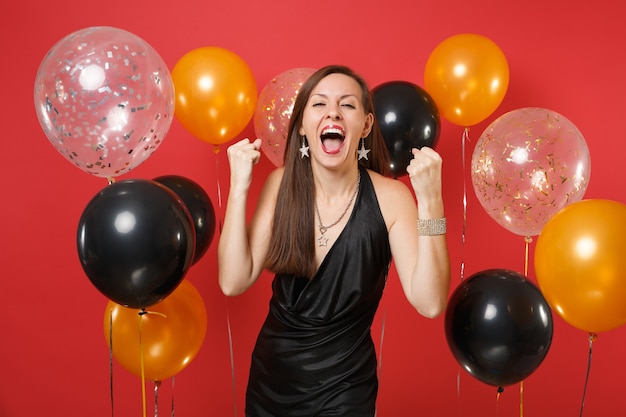 Happy woman in black dress celebrating screaming, clenching fists like winner on bright red background air balloons. International Women's Day, Happy New Year, birthday mockup holiday party concept.