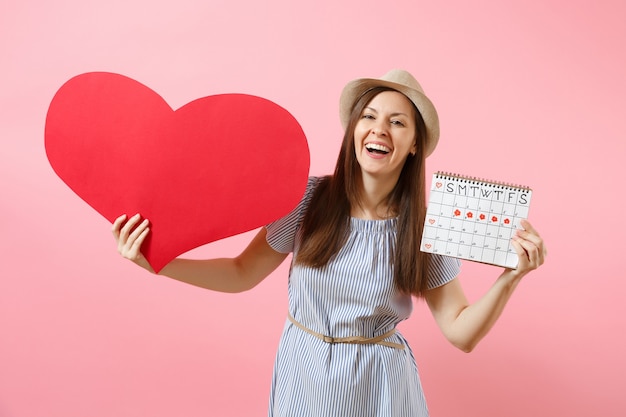 Happy woman in blue dress summer hat holding empty blank big red heart, female periods calendar, checking menstruation days isolated on background. Medical healthcare gynecological concept. Copy space