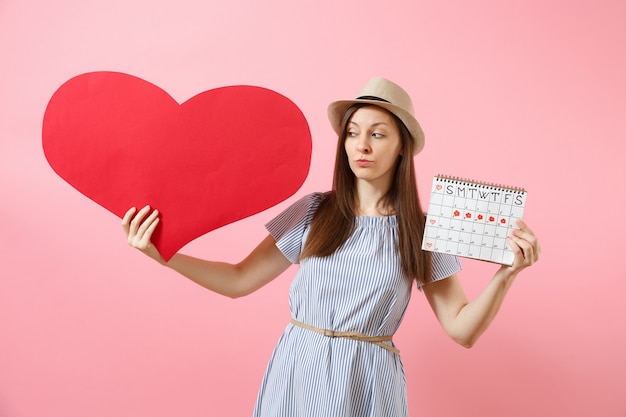 Happy woman in blue dress summer hat holding empty blank big red heart, female periods calendar, checking menstruation days isolated on background. Medical healthcare gynecological concept. Copy space