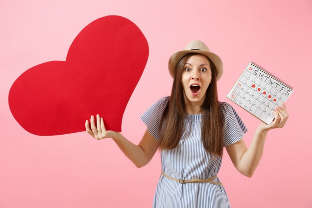 happy woman in blue dress summer hat holding empty blank big red heart, female periods calendar, checking menstruation days isolated on background. medical healthcare gynecological concept. copy space