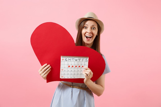 Happy woman in blue dress summer hat holding empty blank big red heart, female periods calendar, checking menstruation days isolated on background. Medical healthcare gynecological concept. Copy space