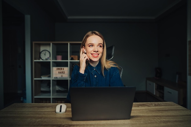 Happy woman in casual clothes sitting at home at a table with a laptop in wireless headphones