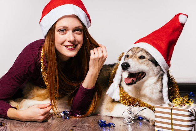クリスマスの帽子をかぶった幸せな女性は犬の隣に横たわっています