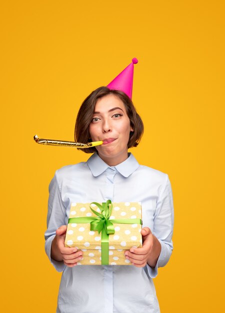 Happy woman congratulating on birthday giving present and blowing party horn looking at camera on yellow background