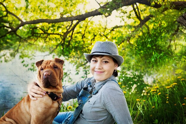 Photo happy woman in denim overalls and hat with her dog shar pei sitting in the meadow near the lake at s