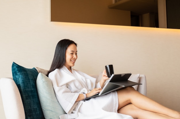 Happy woman in dressing gown sit on couch holding coffee working on laptop.