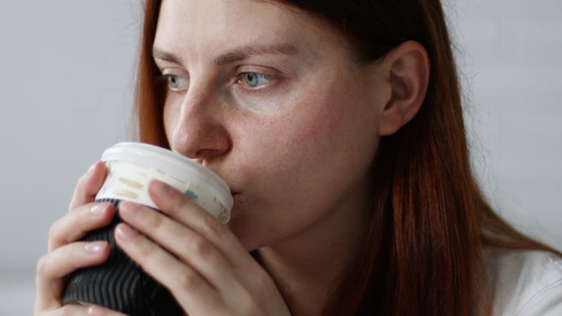 Donna felice che beve caffè dal bicchiere di carta al mattino seduto sul divano di casa