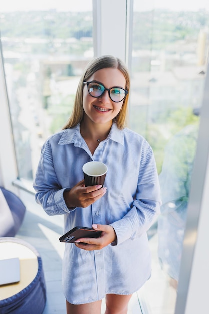 Happy woman drinks coffee and looks out the window in a cafe A young smiling girl in glasses is standing by the window Recreation leisure and free time The modern freelancer lifestyle