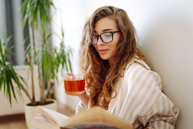 행복한 여성은 뜨거운 음료를 마시고 전화기에 앉아 책을 읽고 잡지를 뒤집습니다.