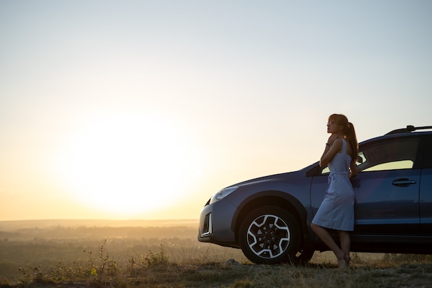 彼女の車の近くで暖かい夜を楽しんでいる青い夏のドレスで幸せな女性ドライバー。旅行と休暇のコンセプト。