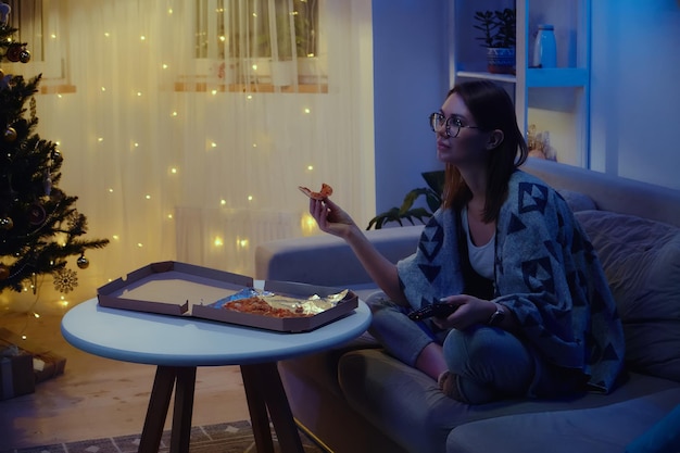 Happy woman eating pizza alone while watching tv at christmas on christmas tree background