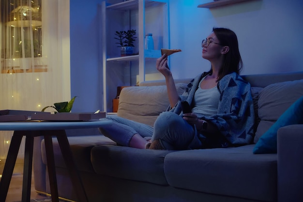 Happy woman eating pizza at night watching tv while sitting on sofa at home