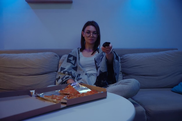 Happy woman eating pizza at night watching tv while sitting on\
sofa at home