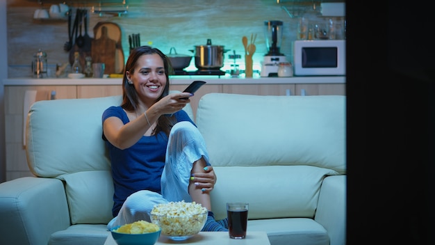 Photo happy woman eating popcorn on sofa and watching tv in living room at home. excited, amused, lonely lady enjoying the evening sitting on comfortable couch dressed in pajamas in front of television.