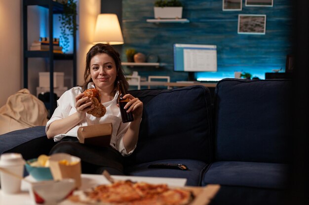 Happy woman eating tasty takeaway hamburger holding beer bottle relaxing on couch watching television family sitcom. Smiling person having takeout burger and fast food delivery menu.