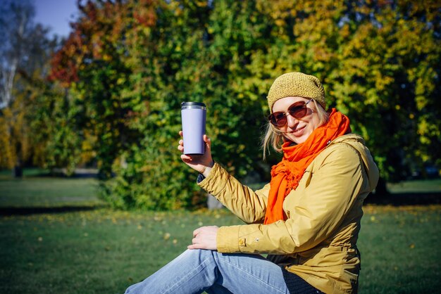 緑の葉の背景に公園のベンチでコーヒーを楽しんで幸せな女性。秋のコンセプト。晴れた秋の日の余暇。スペースをコピーします。