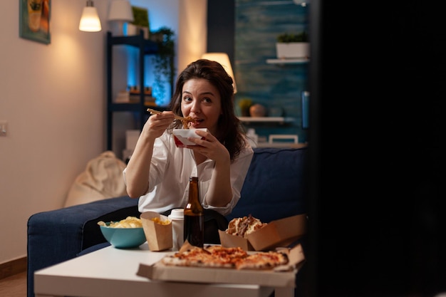 Happy woman enjoying delicious unhealthy takeaway asian noodles
with chopsticks while watching television in home living room.
person sitting on couch eating chinese ramen box and junk
food.