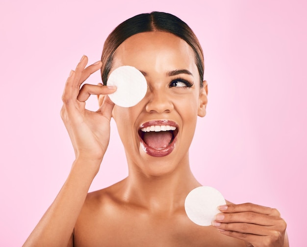 Happy woman face and cotton pad in beauty makeup removal or skincare cosmetics against a pink studio background Excited female person or model with cosmetic swab for cleaning or facial treatment