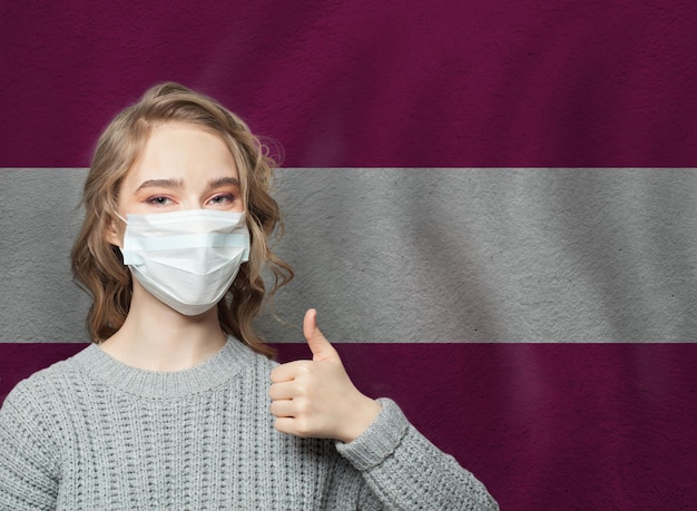 Foto donna felice con una maschera che tiene il pollice in alto sullo sfondo della bandiera della lettonia concept di epidemia di influenza e protezione dal virus