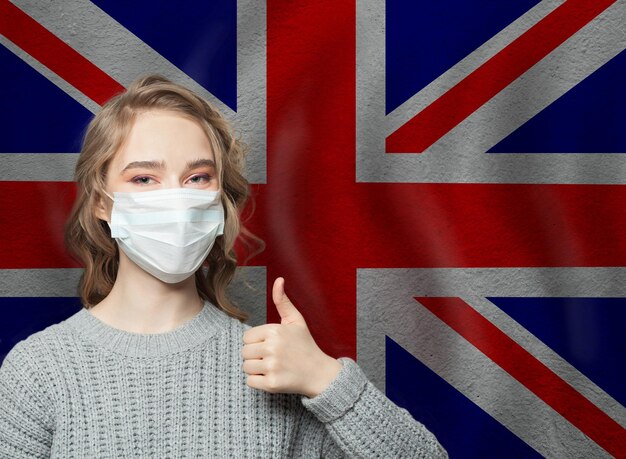 Happy woman in face mask holding thumb up on national flag UK background Flu epidemic and virus protection concept