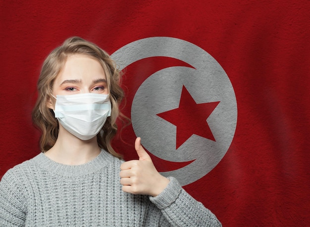 Happy woman in face mask holding thumb up on Tunisia flag background Flu epidemic and virus protection concept