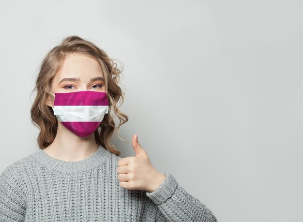 Foto donna felice con la maschera facciale che tiene il pollice in alto con la bandiera della latvia sullo sfondo concept di protezione contro l'epidemia di influenza e il virus