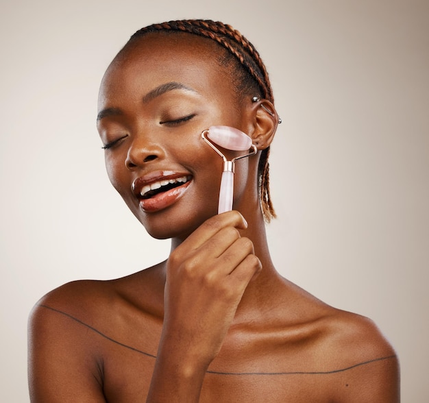 Happy woman face roller and massage for beauty rose quartz cosmetics and aesthetic dermatology in studio on brown background African model crystal stone and lymphatic drainage for facial skincare