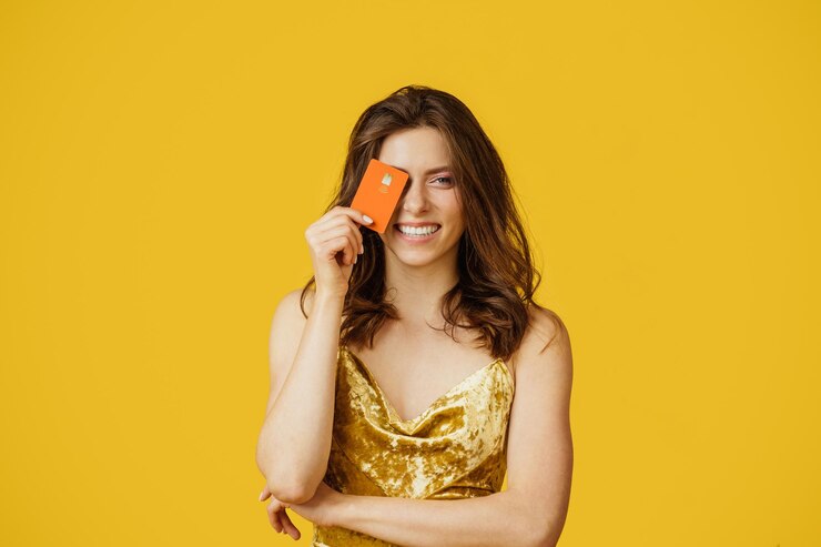 Photo happy woman in festive dress holding credit card near eye and smiling at camera isolated on yellow background