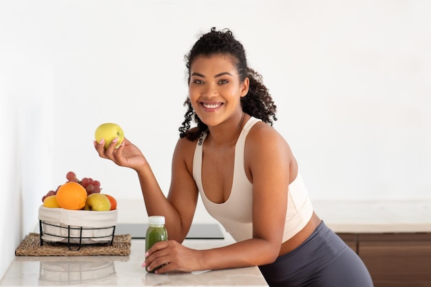 Happy woman in fitwear holding apple and smoothie in kitchen