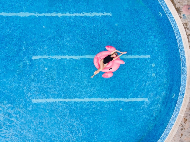 Happy woman on flamingo pool float in pool, drone aerial view