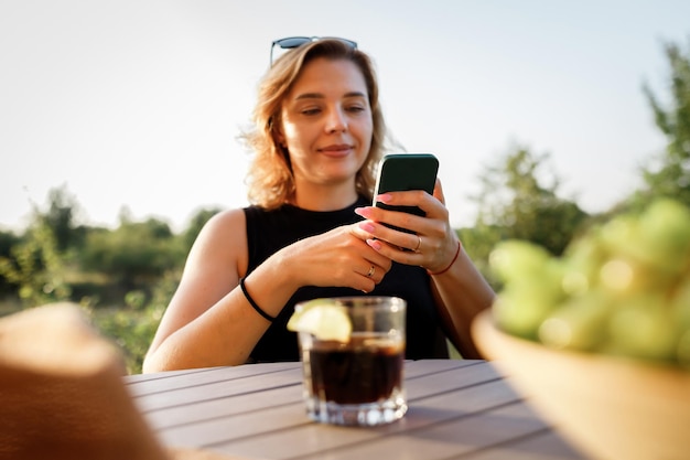 Happy Woman freelancer gebruikt telefoon en drinkt cocktail terwijl u ontspant op gezellige glamping-tent op zonnige dag Luxe kampeertent voor buiten zomervakantie en vakantie Lifestyle concept