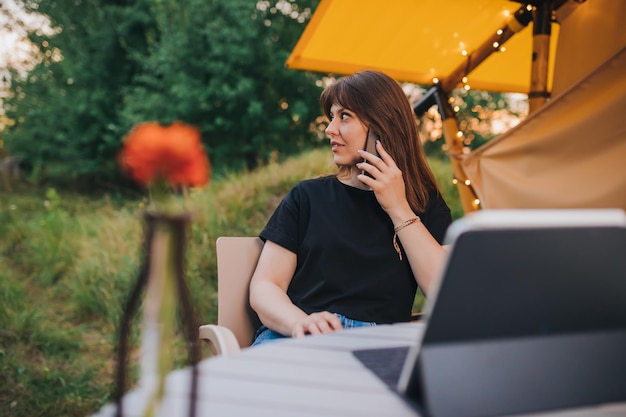 Happy woman freelancer talking phone and using laptop on cozy\
glamping tent in a sunny day luxury camping tent for outdoor summer\
holiday and vacation lifestyle concept