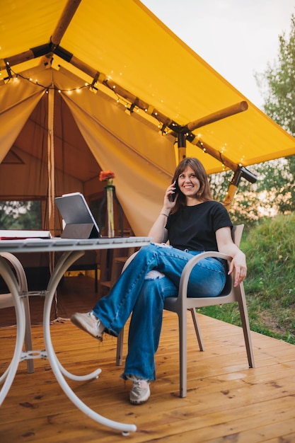 Happy Woman freelancer talking phone and using laptop on cozy glamping tent in a sunny day Luxury camping tent for outdoor summer holiday and vacation Lifestyle concept