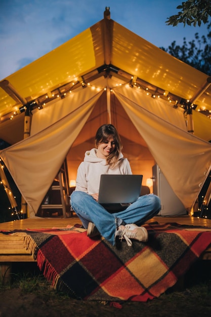 Happy Woman freelancer using a laptop on a cozy glamping tent in a summer evening Luxury camping tent for outdoor holiday and vacation Lifestyle concept