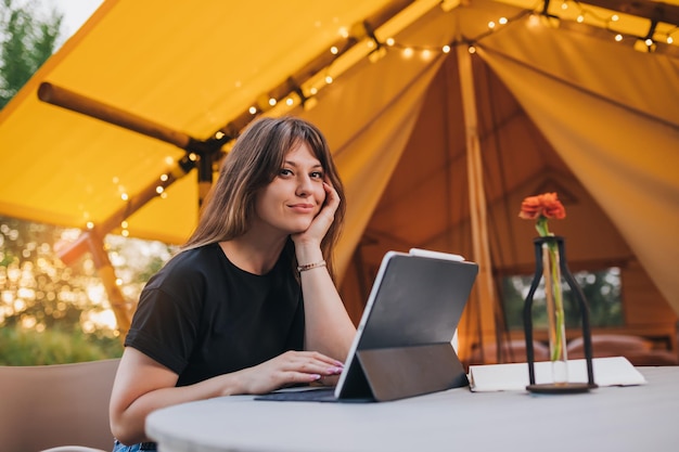 Happy Woman freelancer using a laptop on a cozy glamping tent in a sunny day Luxury camping tent for outdoor summer holiday and vacation Lifestyle concept