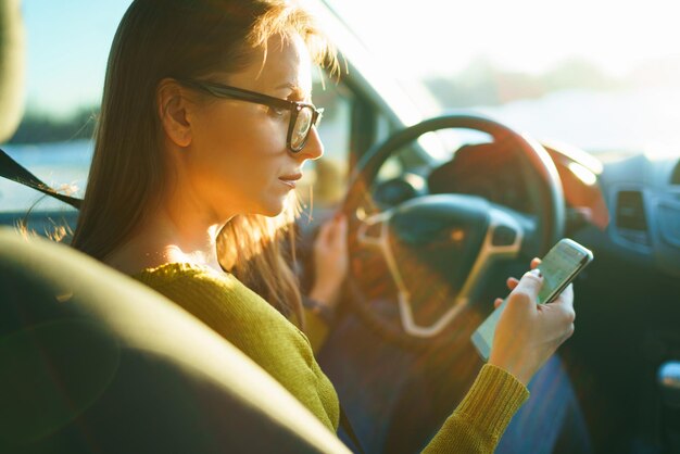 Happy woman in glasses uses a smartphone while driving a car at sunset