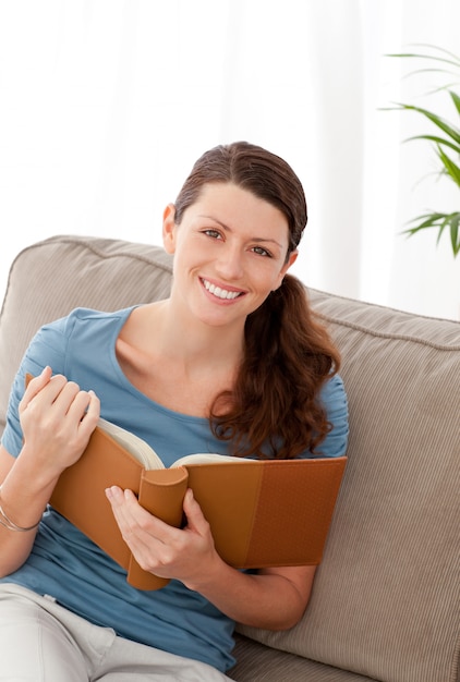 Happy woman holding a book sitting on her sofa