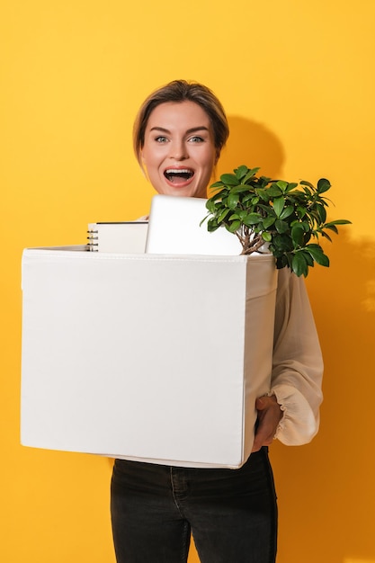 Happy woman holding box with personal items after job promotion against yellow background