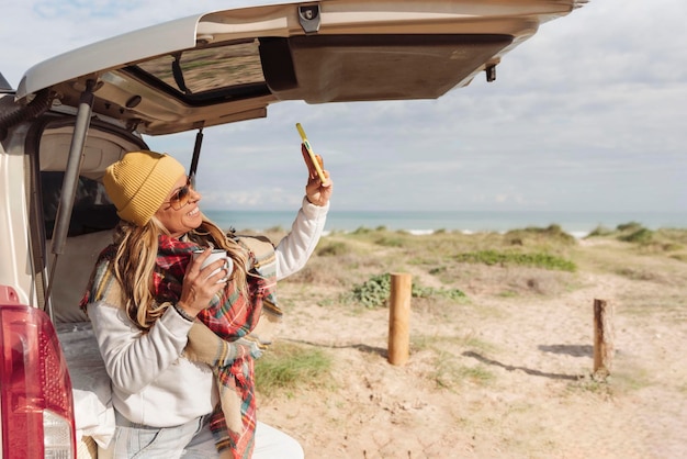 Happy woman holding a mug leaning against a van next to beach and taking a selfie