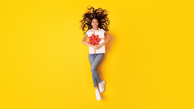 Happy woman holding wrapped present box over yellow studio background