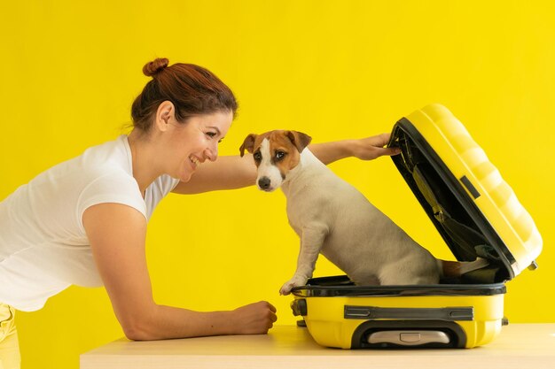 Happy woman holds an open suitcase with a dog inside on a yellow background