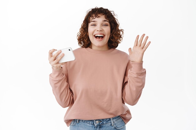 Happy woman laughing, holding smartphone and celebrating, triumphing, winning on mobile video game, standing cheerful against white background