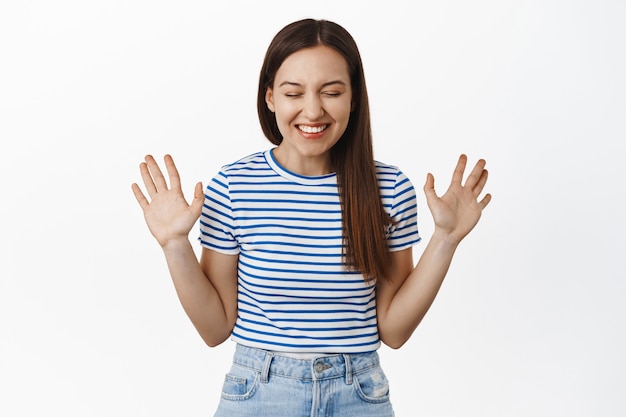 Happy woman laughing, smiling white teeth, raising empty hands up, standing cheerful and excited, wearing striped summer t-shirt and jeans, white wall