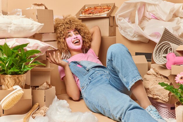 Happy woman lies around carton packages looks above applies
beauty mask dressed in denim overalls glad to move in first home
tires due to long relocation hard difficult moving day and
eviction